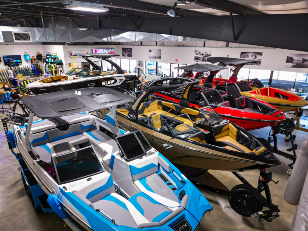 Interior view of a boat showroom with a variety of boats on display, featuring different sizes and colors, including blue and white, red, and tan. Wakeboard towers, bimini tops, and sleek designs are prominent. The showroom is well-lit and includes racks of water sports equipment in the background.